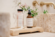 Two bottles of Ivy & Sage products on a wooden surface next to sinks in bathroom with a blurred background