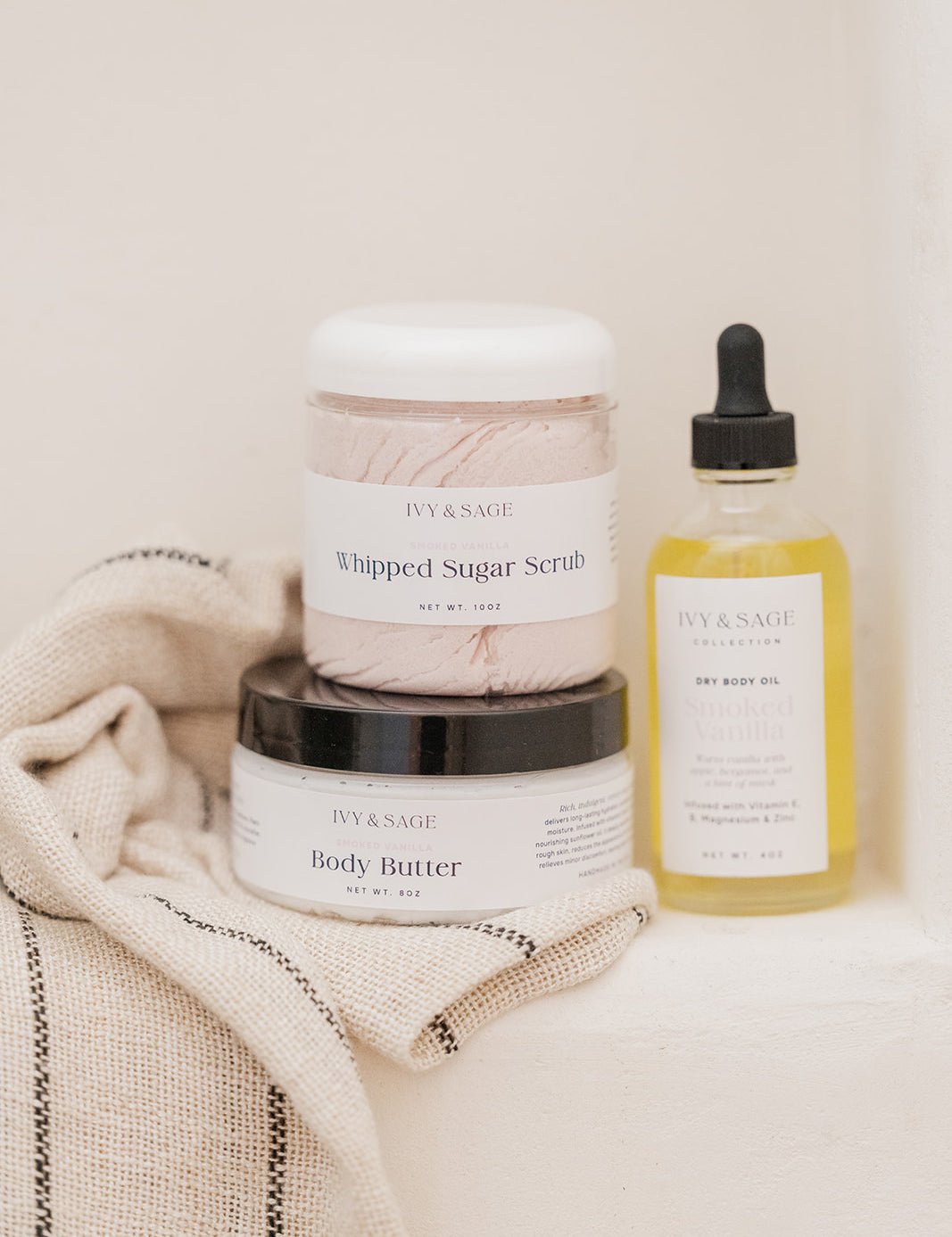 IVY & SAGE skincare products including a jar of body butter, a jar of whipped sugar scrub, and a bottle of body oil on a textured surface.