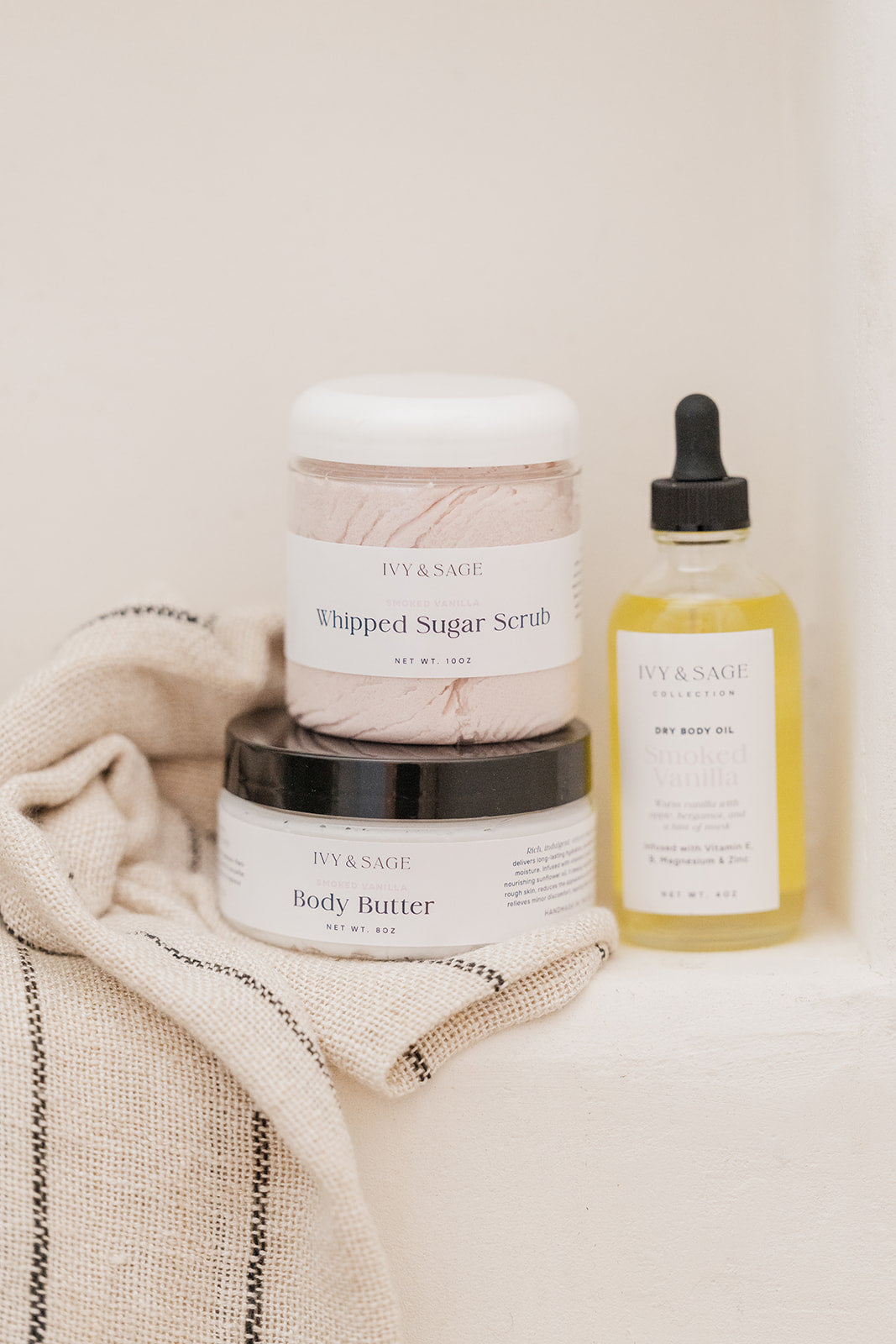 IVY & SAGE skincare products including a jar of body butter, a jar of whipped sugar scrub, and a bottle of body oil on a textured surface.