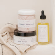 IVY & SAGE skincare products including a jar of body butter, a jar of whipped sugar scrub, and a bottle of body oil on a textured surface.