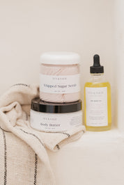 IVY & SAGE skincare products including a jar of body butter, a jar of whipped sugar scrub, and a bottle of body oil on a textured surface.