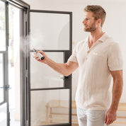 Man in a white shirt and shorts standing near a glass door, holding and spraying Room & Linen Spray