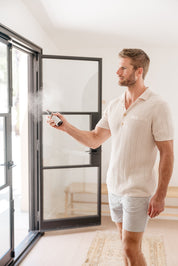 Man in a white shirt and shorts standing near a glass door, holding and spraying Room & Linen Spray