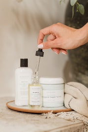 Hand holding a dropper over a bottle of skincare product on a wooden tray with other skincare items.