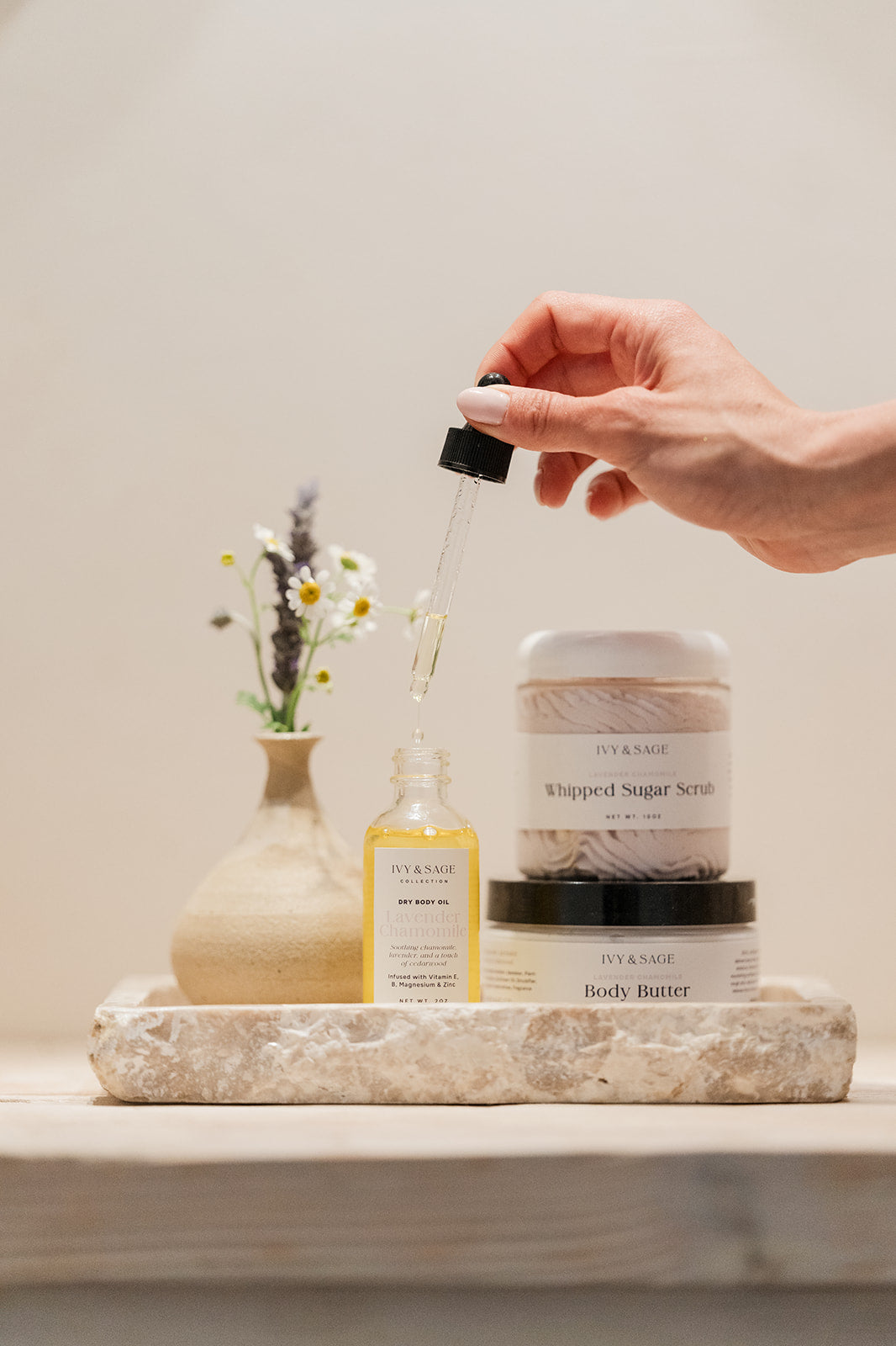 Hand holding a body oil dropper with a jar of body butter and sugar scrub with a neutral background