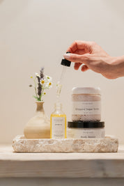 Hand holding a body oil dropper with a jar of body butter and sugar scrub with a neutral background