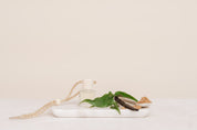Glass Car Diffuser bottle with a string on a white marble slab with green leaves and a piece of wood against a beige background for Caramelised Sugar