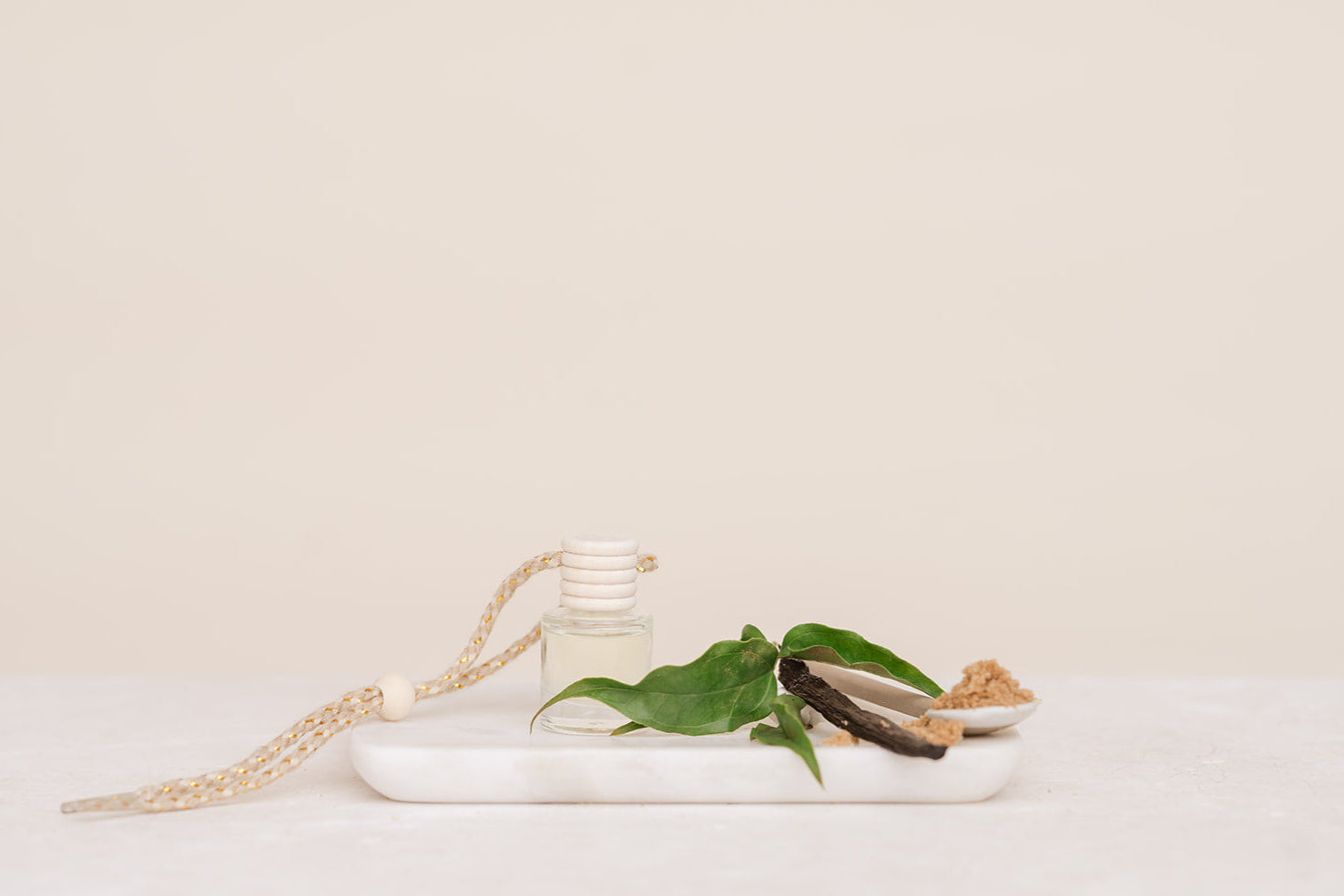 Glass Car Diffuser bottle with a string on a white marble slab with green leaves and a piece of wood against a beige background for Caramelised Sugar