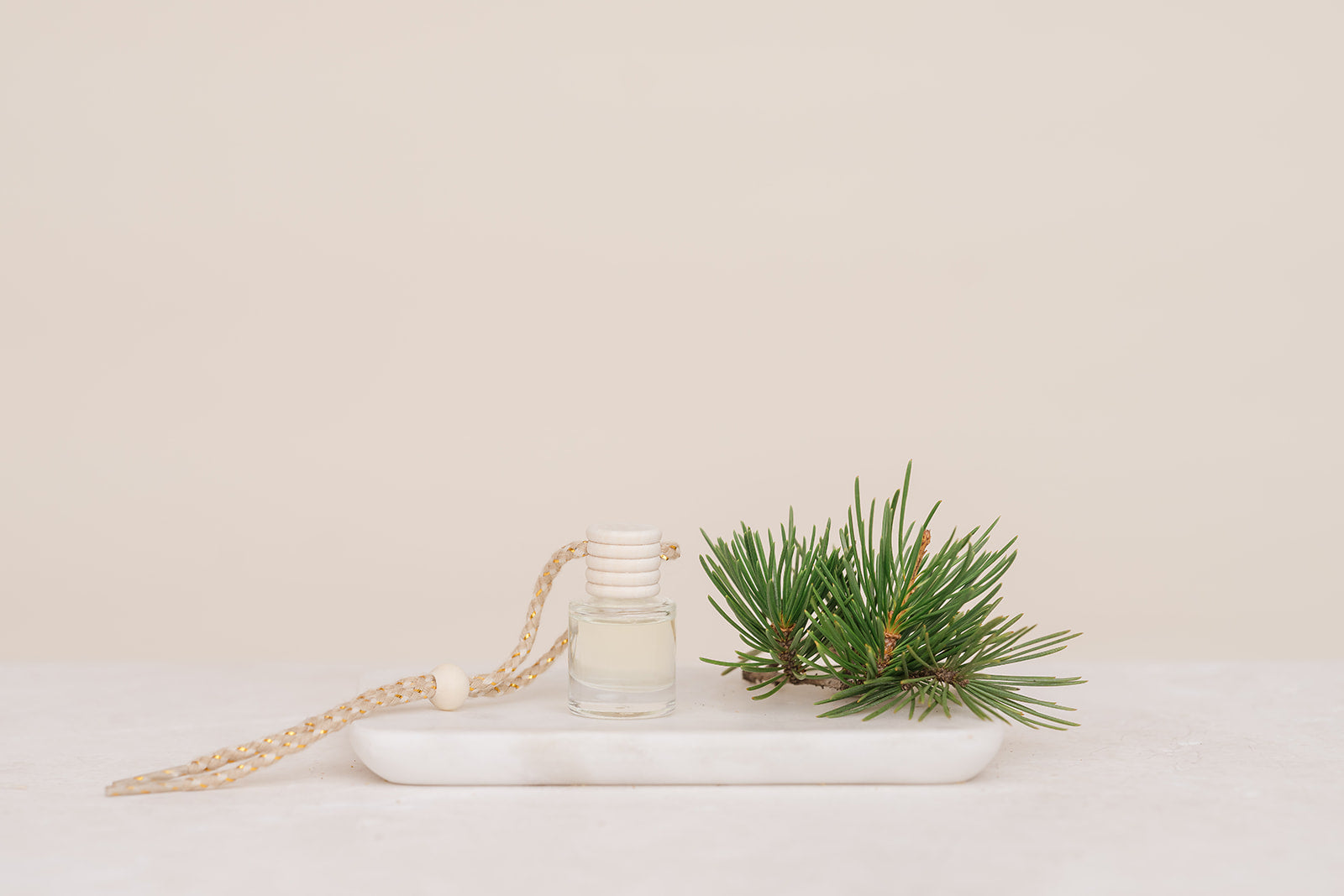 Small glass car diffuser with an adjustable rope and a bundle of pine needles on a white marble slab against a beige background. Car Freshener that smells like Christmas. Christmas scents perfect gift idea. 