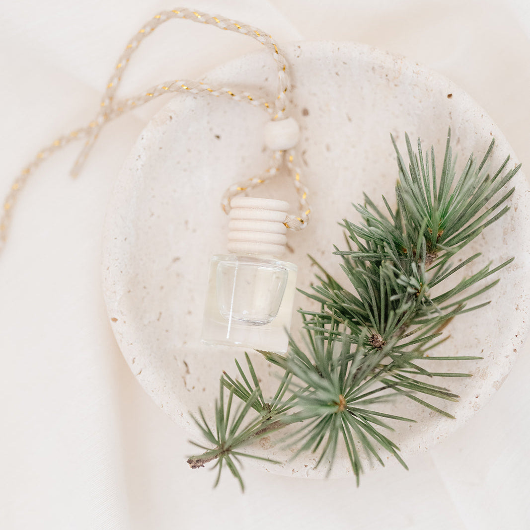 Small glass car diffuser with an adjustable rope and a bundle of pine needles on a white marble slab against a beige background. Car Freshener that smells like Christmas. Christmas scents perfect gift idea. 