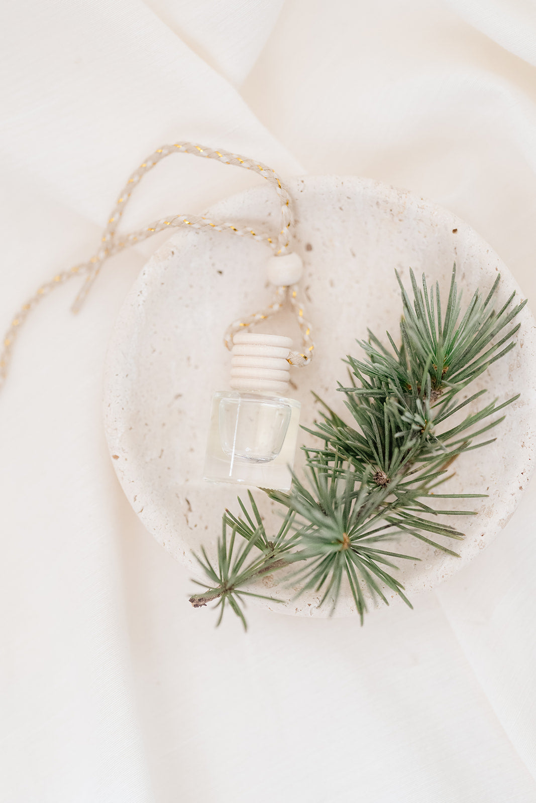 Small glass car diffuser with an adjustable rope and a bundle of pine needles on a white marble slab against a beige background. Car Freshener that smells like Christmas. Christmas scents perfect gift idea. 