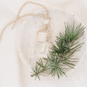 Small glass car diffuser with an adjustable rope and a bundle of pine needles on a white marble slab against a beige background. Car Freshener that smells like Christmas. Christmas scents perfect gift idea. 