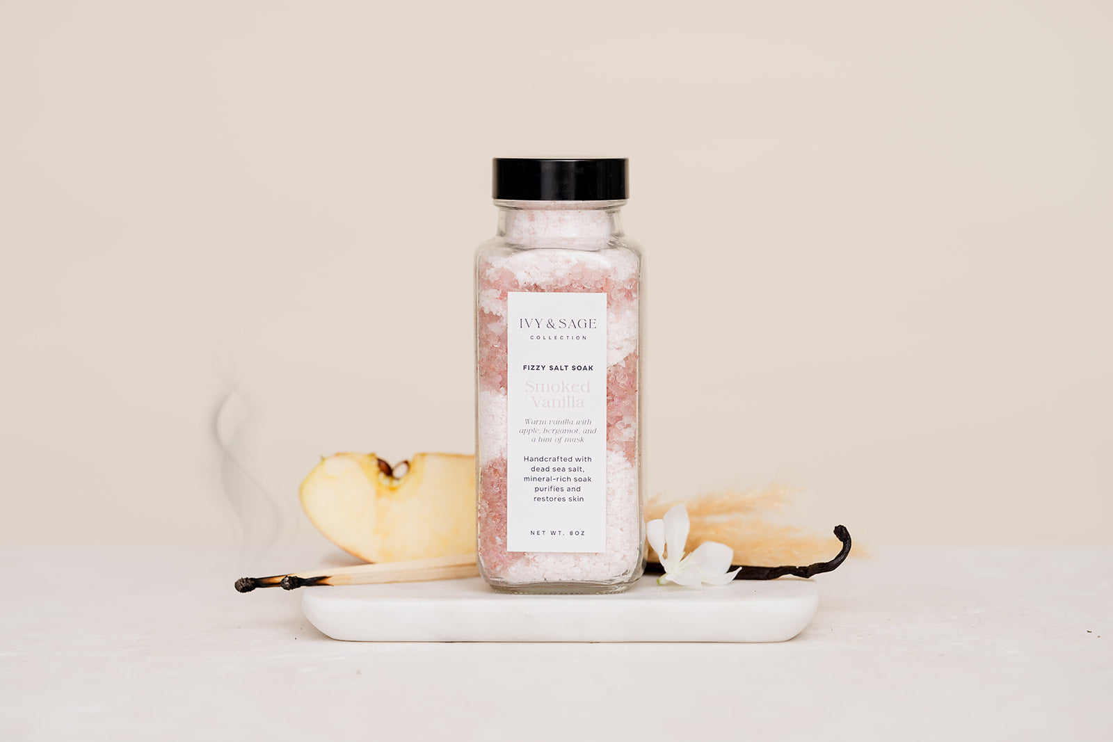 Bottle of Salt Soak with a label on a white surface with vanilla beans and a spoon.