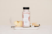 Bottle of Salt Soak with a label on a white surface with vanilla beans and a spoon.