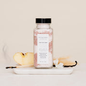 Bottle of Salt Soak with a label on a white surface with vanilla beans and a spoon.