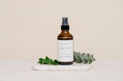 Bottle of Room & Linen Spray on a white tray with green leaves on a beige background