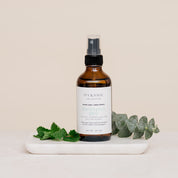 Bottle of Room & Linen Spray on a white tray with green leaves on a beige background