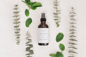 Bottle of Room & Linen Spray surrounded by eucalyptus leaves on a white background