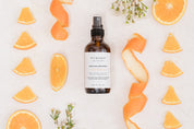 Bottle of Room & Linen Spray surrounded by orange slices and peels on a light background