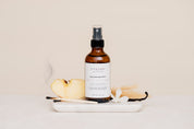 Bottle of Ivy & Sage Room & Linen Spray on a white plate with apple, vanilla and a smokey match