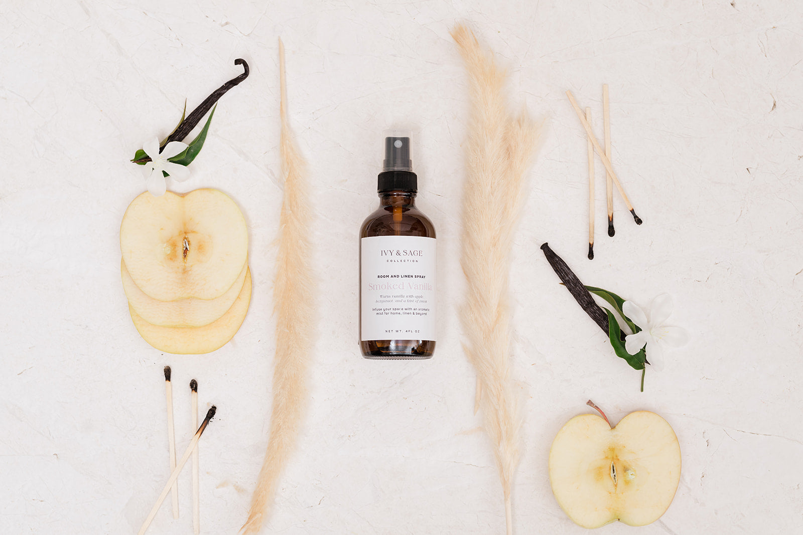 A bottle of Room & Linen Spray surrounded by apples, vanilla beans, and reeds on a light background