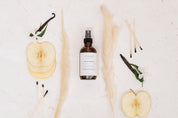A bottle of Room & Linen Spray surrounded by apples, vanilla beans, and reeds on a light background