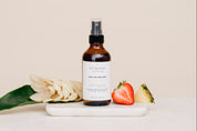 Bottle of Room & Linen Spray on a white surface with flowers, strawberry and pinappe