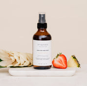 Bottle of Room & Linen Spray on a white surface with flowers, strawberry and pinappe