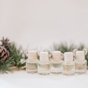 Small glass car diffuser with an adjustable rope and a bundle of pine needles on a white marble slab against a beige background. Car Freshener that smells like Christmas. Christmas scents perfect gift idea. 