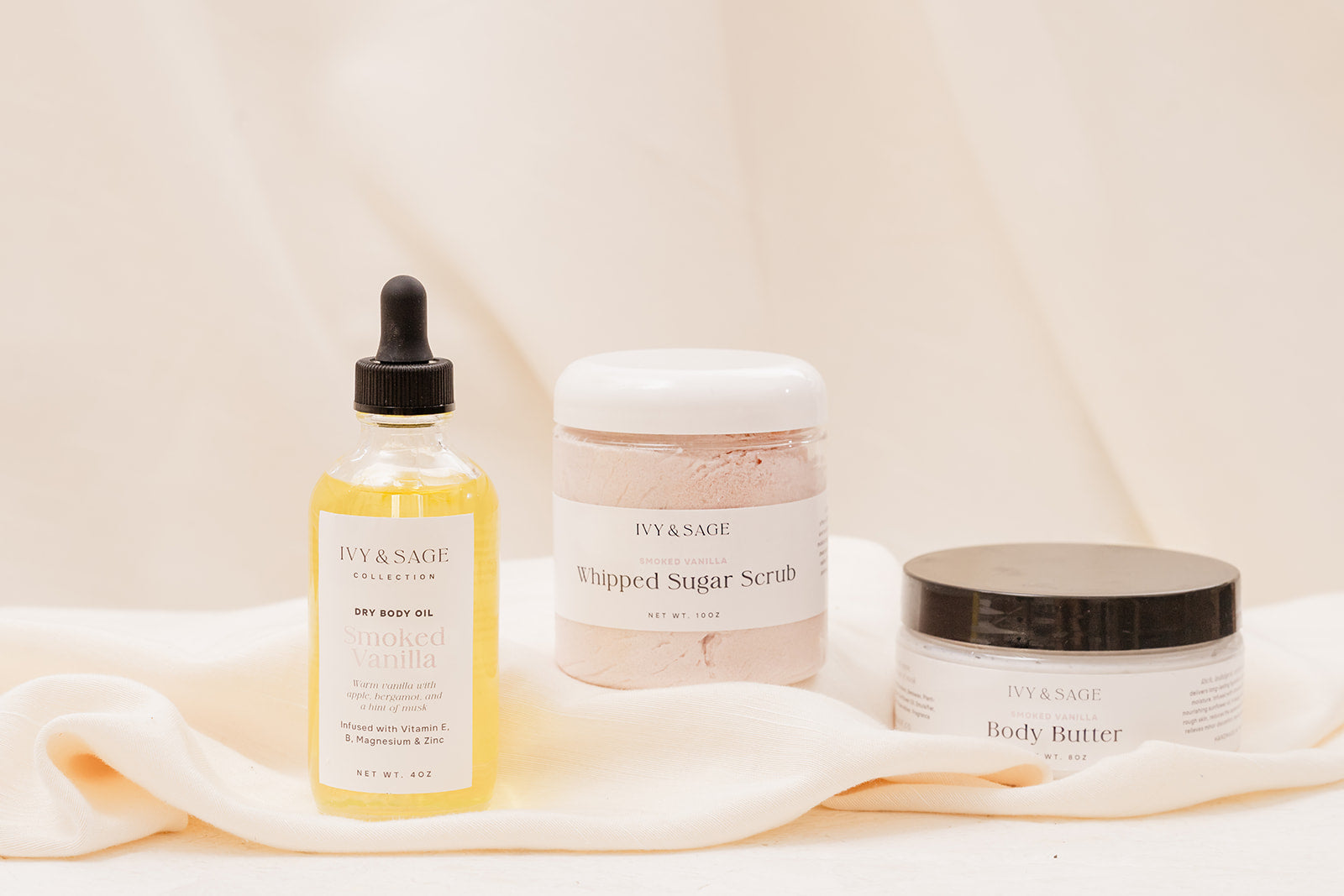 IVY & SAGE skincare products including a bottle of oil and body butter and sugar scrubs on a soft fabric background.
