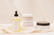 IVY & SAGE skincare products including a bottle of oil and body butter and sugar scrubs on a soft fabric background.