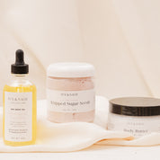 IVY & SAGE skincare products including a bottle of oil and body butter and sugar scrubs on a soft fabric background.